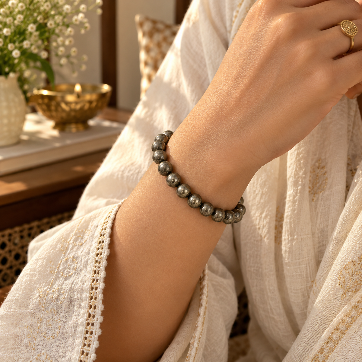 A close-up of a person's wrist wearing a beaded bracelet made of polished dark gray stone beads. The person is wearing a cream-colored embroidered robe, and a gold ring is visible on their finger.