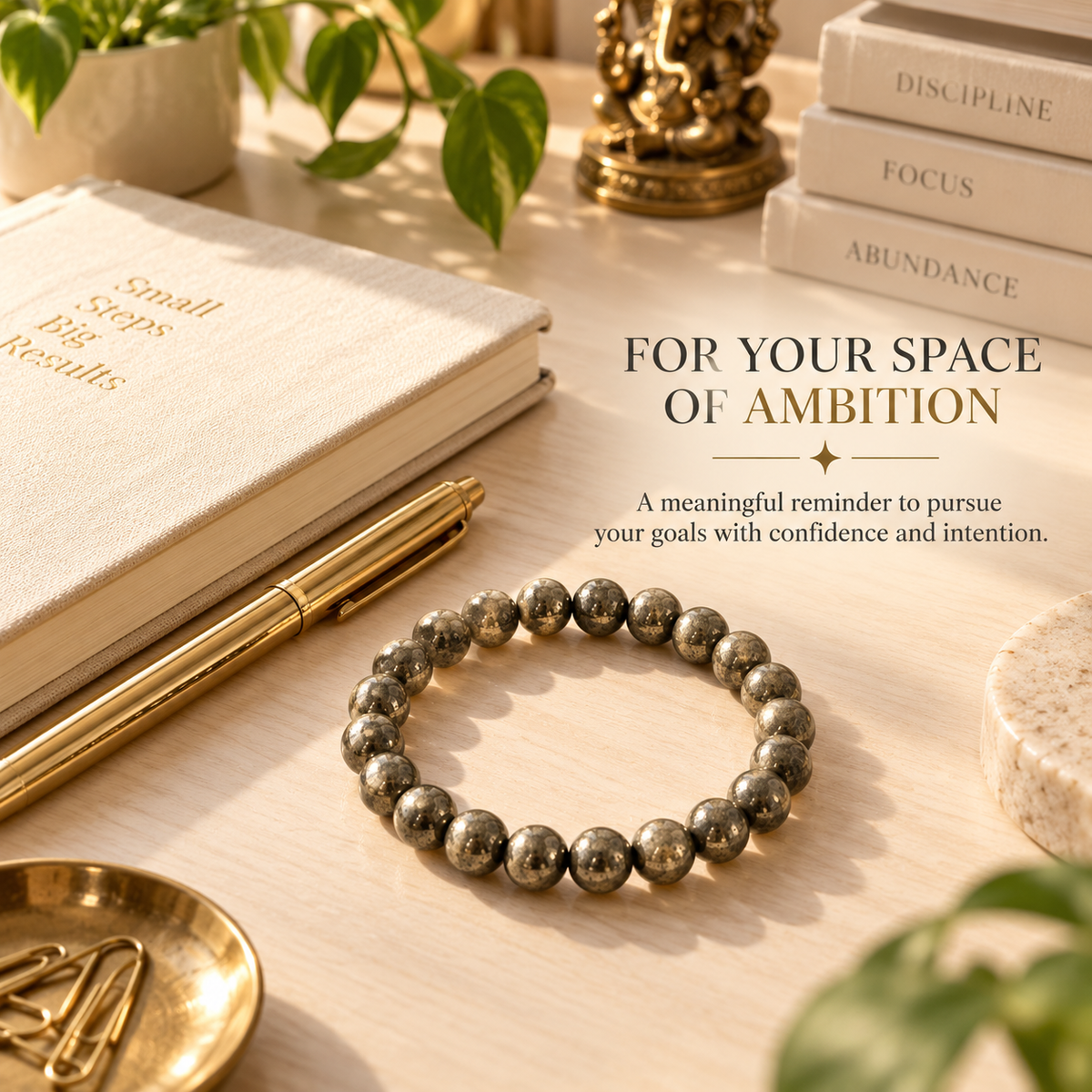 A beaded bracelet made of metallic gray round beads is displayed on a light wood desk alongside a cream notebook, gold pen, books, and decorative objects. Text on the image reads 'FOR YOUR SPACE OF AMBITION'.