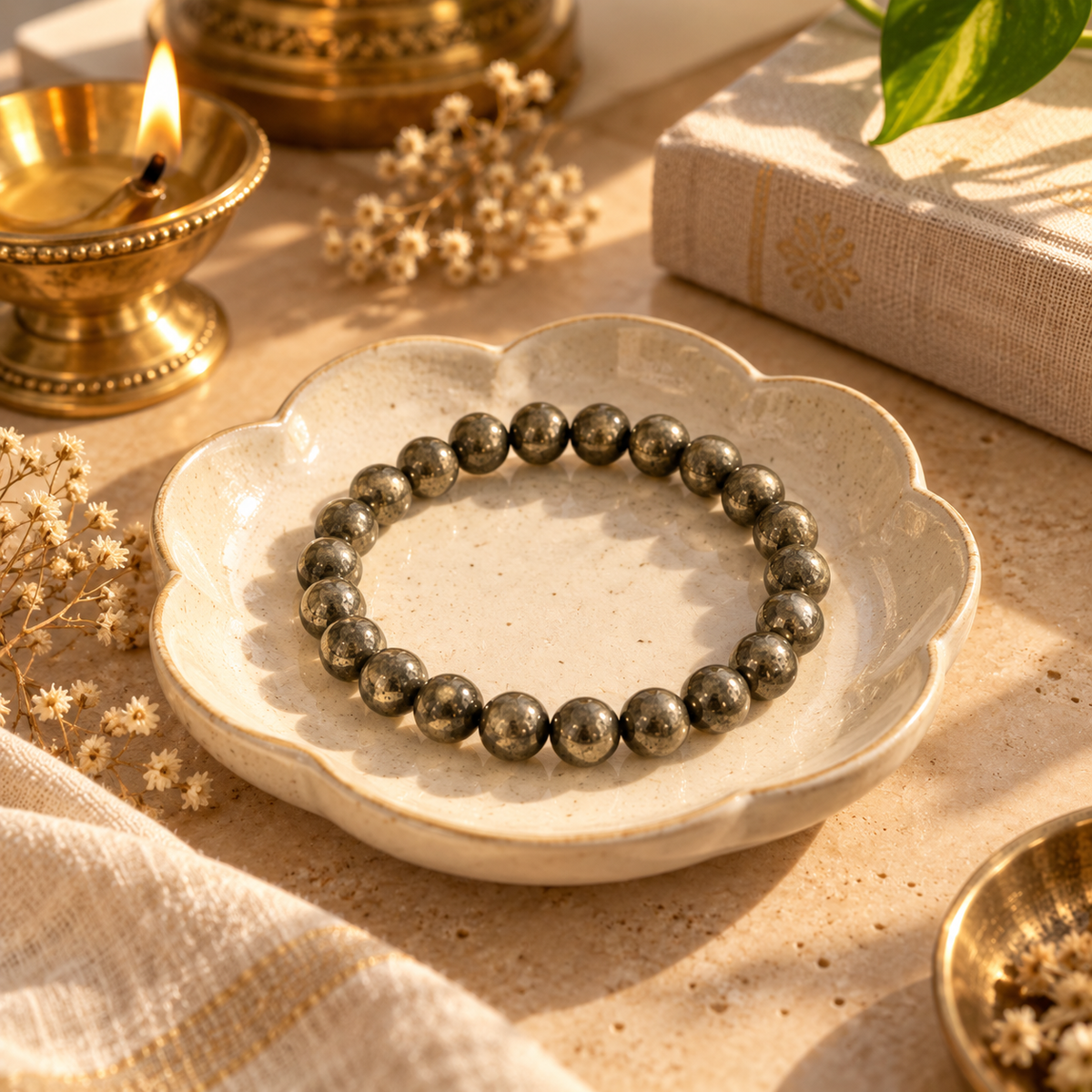 A beaded bracelet made of polished metallic gray spheres, displayed on a scalloped cream ceramic dish. The scene is styled with warm, golden lighting, featuring a brass oil lamp, dried flowers, a linen book, and a green leaf in the background.