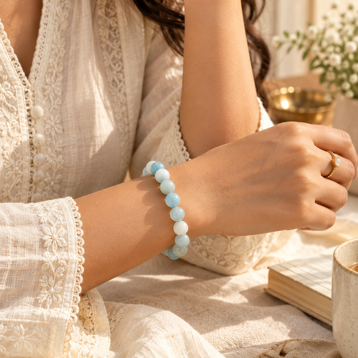 A person wearing a light blue beaded bracelet on their wrist, dressed in a cream lace blouse, with a warm, softly lit background featuring a book and a cup.