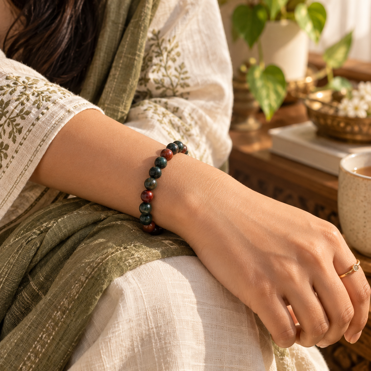 A close-up lifestyle shot of a person's wrist wearing a beaded bracelet made of dark green, red, and black beads, styled with an embroidered cream dress and a green shawl in a warm, sunlit indoor setting.