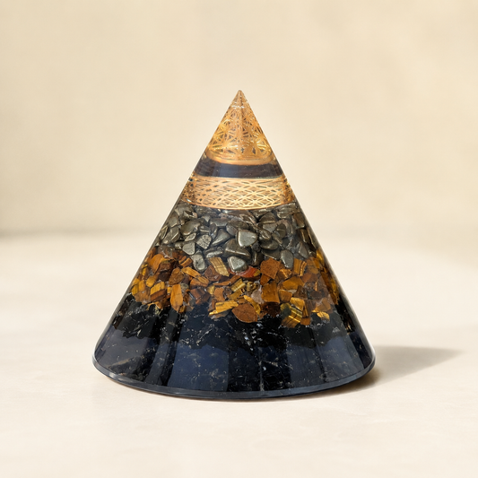 A conical crystal tower or chakra ornament made of layered black and gray stone with embedded gold and brown metallic flakes, topped with a gold-patterned pointed cap, photographed on a warm beige background.
