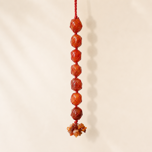 A vertical hanging ornament made of seven faceted orange-red beads on a red cord, with a small cluster of polished stone beads at the bottom, shown against a plain warm beige background.