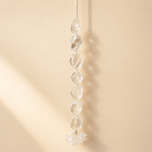 A clear faceted crystal suncatcher with seven stacked round beads and a pointed bottom, hanging against a warm beige wall with soft sunlight and shadows.