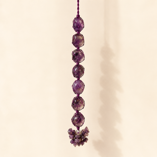 A hanging amethyst crystal ornament with seven faceted purple beads and a small cluster of amethyst stones at the bottom, shown against a warm beige background.