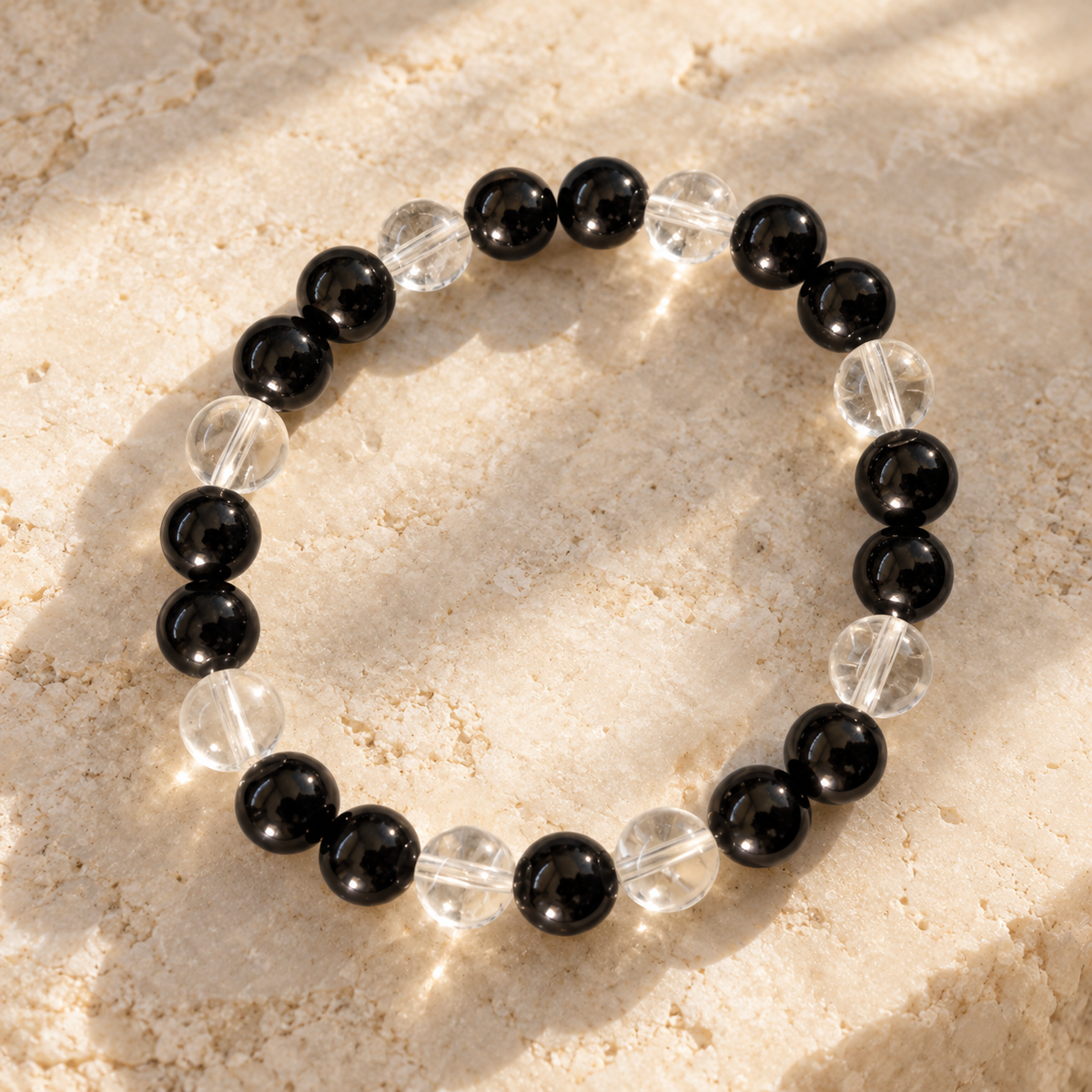 A beaded stretch bracelet featuring alternating glossy black and clear faceted beads, arranged in a circle on a textured beige stone surface with soft sunlight and shadows.