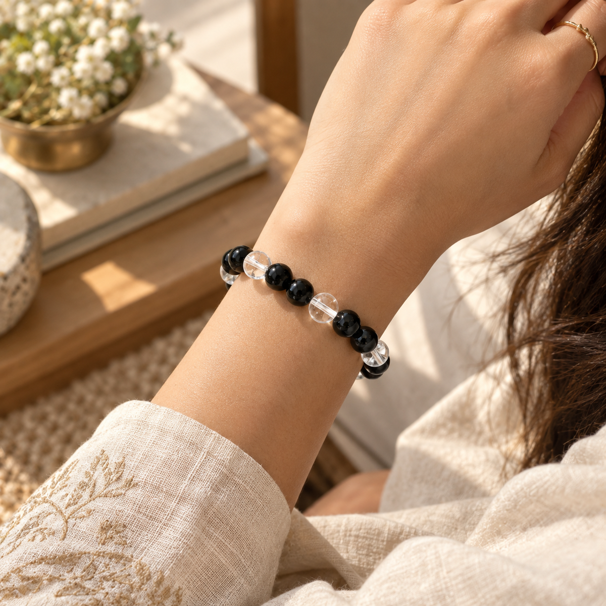 A close-up of a person's wrist wearing a beaded bracelet made of alternating black and clear faceted beads, styled with a cream embroidered sleeve and a gold ring on another finger.