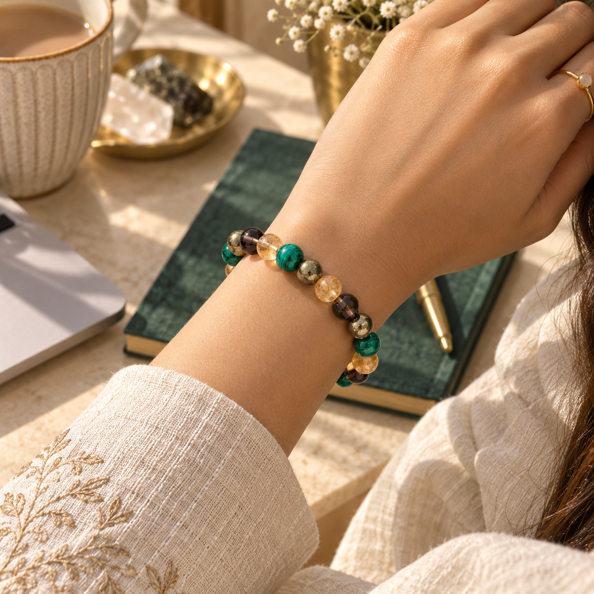 A close-up lifestyle shot of a person's wrist wearing a beaded bracelet made of green, amber, and metallic beads, set against a cozy desk background with a notebook, coffee cup, and ring.