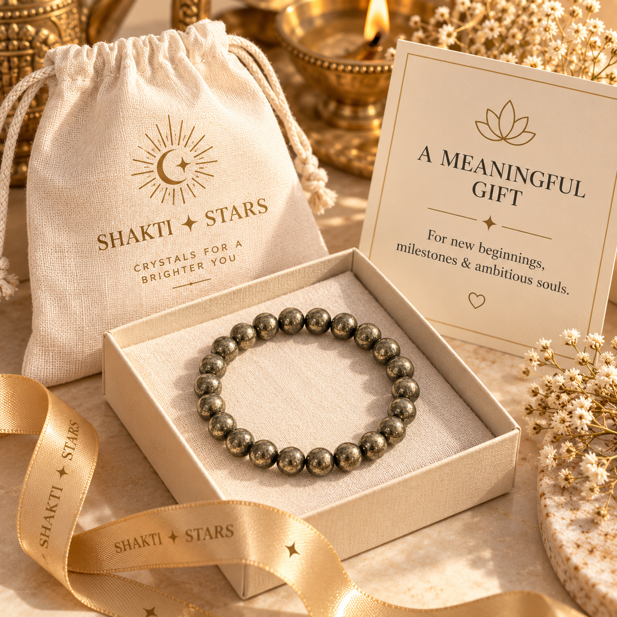 A beaded bracelet made of polished metallic gray-green stones sits in an open cream gift box, surrounded by a branded cream drawstring pouch, a gift card reading 'A MEANINGFUL GIFT,' and a gold ribbon. The visible brand on the packaging is Shakti Stars.