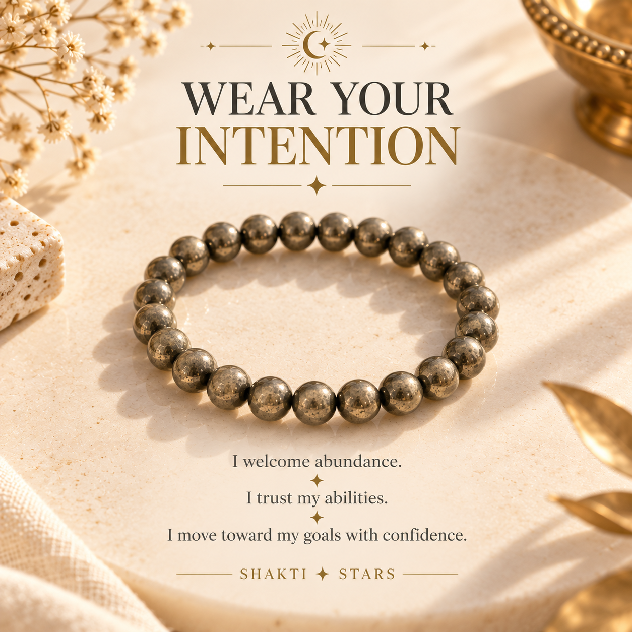 A metallic beaded stretch bracelet made of dark gold-toned round beads, displayed on a textured beige surface with decorative dried flowers and gold-toned props. The image includes the text 'WEAR YOUR INTENTION' and 'SHAKTI STARS'.