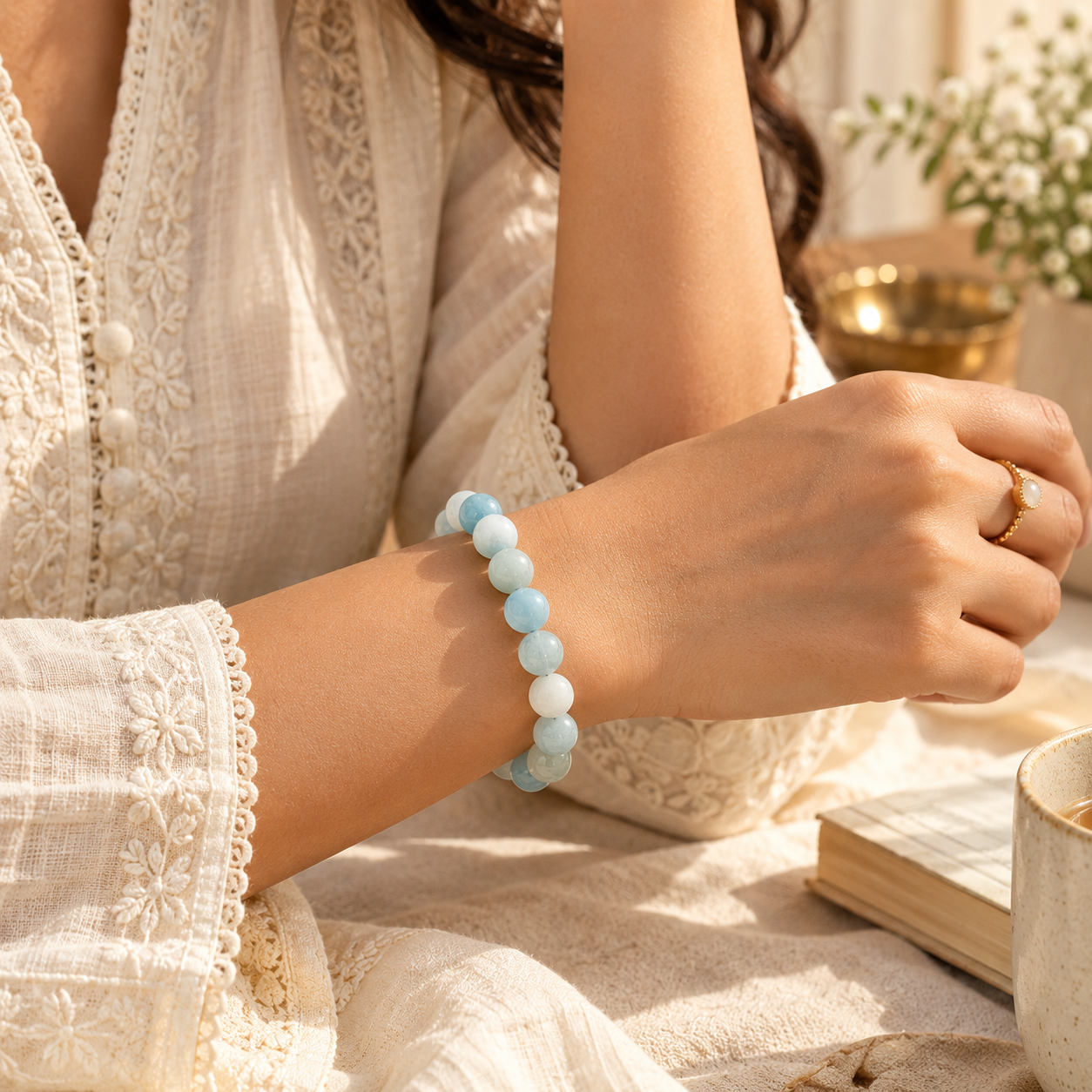A person wearing a light blue beaded bracelet on their wrist, dressed in a cream lace blouse, with a warm, softly lit background featuring a book and a cup.