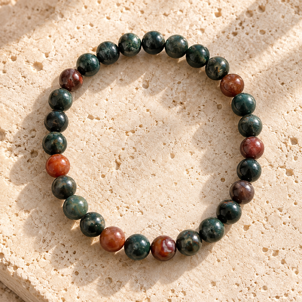 A beaded stretch bracelet made of dark green and reddish-brown polished stone beads, arranged in a circle on a textured beige stone surface.