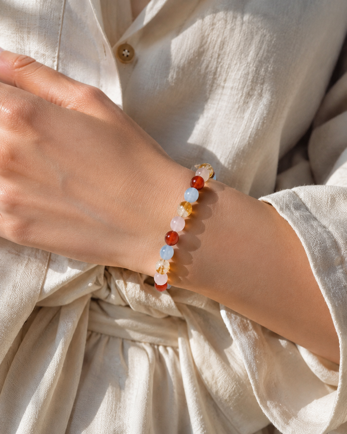 A close-up of a person's wrist wearing a delicate beaded bracelet, styled with a cream linen shirt in warm natural sunlight.